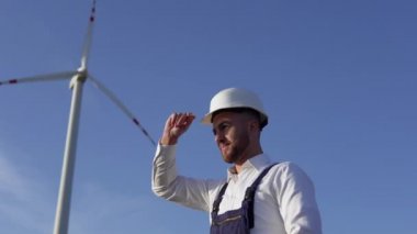 Elektrik santrali mühendisi beyaz klasik bir gömlek, mavi tulum ve yel değirmeni arka planında beyaz bir miğfer giyiyor.