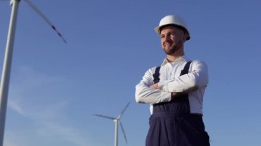 Elektrik santrali mühendisi beyaz klasik bir gömlek, mavi tulum ve yel değirmeni arka planında beyaz bir miğfer giyiyor.