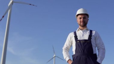Elektrik santrali mühendisi beyaz klasik bir gömlek, mavi tulum ve yel değirmeni arka planında beyaz bir miğfer giyiyor.