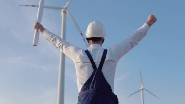 Mühendis elinde projenin çizimlerini içeren büyük bir kağıt tutuyordu. İşçi, arkası kameraya dönük olarak yel değirmenlerinin arkasında durur ve elleri havada sevinir.