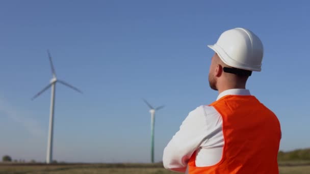 Ein männlicher Ingenieur mit weißem Helm, klassischem Hemd und orangefarbener Arbeitsweste begutachtet die Windräder eines Kraftwerks — Stockvideo