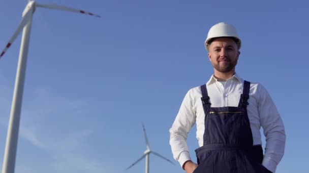 O engenheiro da usina está de pé em uma camisa clássica branca, macacão azul e um capacete branco em um fundo de moinho de vento — Vídeo de Stock
