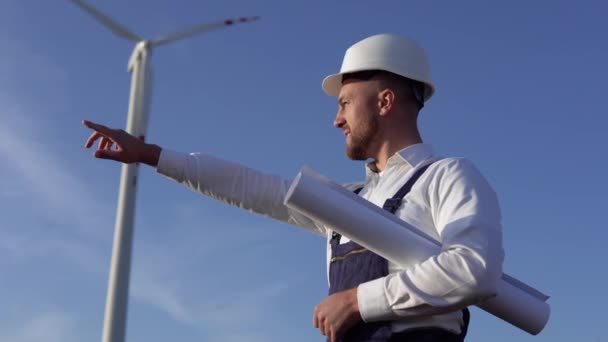 Um engenheiro em um capacete branco, macacão azul de trabalho e uma camisa clássica examina as turbinas eólicas de uma usina — Vídeo de Stock