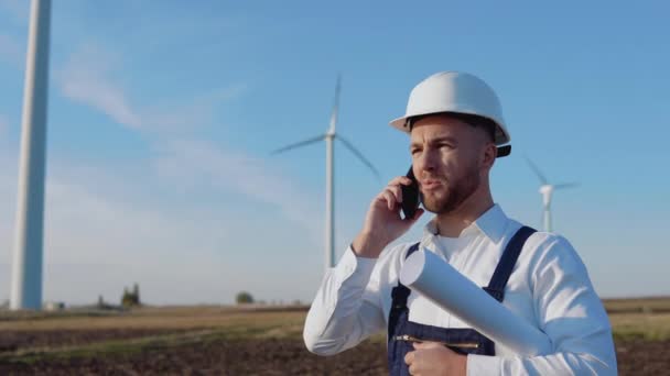Inżynier w białym hełmie, pracujący w niebieskich kombinezonach i w klasycznej koszuli trzyma w ręku duży zwinięty arkusz papieru z rysunkami projektu i rozmawia przez komórkę przy turbinach wiatrowych. — Wideo stockowe