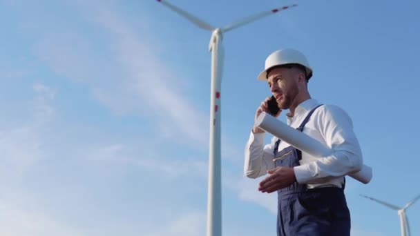 Мужчина-инженер в белом шлеме, работает в синем комбинезоне и в классической рубашке держит в руке большой свернутый лист бумаги с рисунками проекта и разговаривает по мобильному телефону возле ветряных турбин — стоковое видео