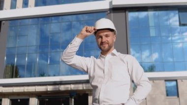 Beyaz gömlekli ve kasklı bir mühendis modern cam bir binanın arka planında duruyor. Modern yapı