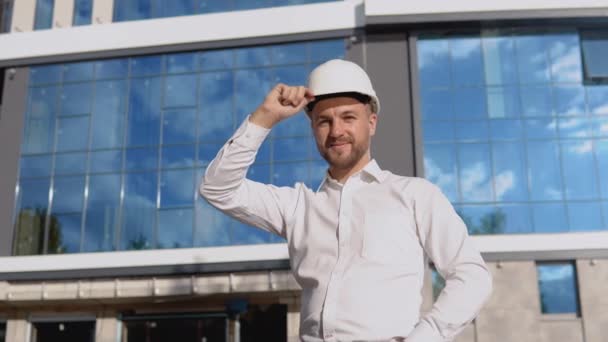 モダンなガラスの建物を背景に、白いシャツとヘルメットのエンジニアが立っています。近代建築 — ストック動画