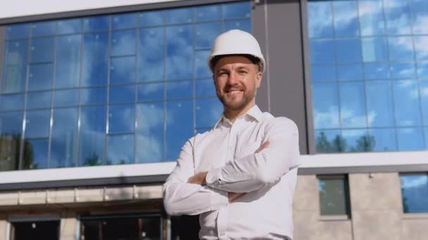 An engineer in a white shirt and helmet stands against the backdrop of a modern glass building. Modern construction — Stock Video