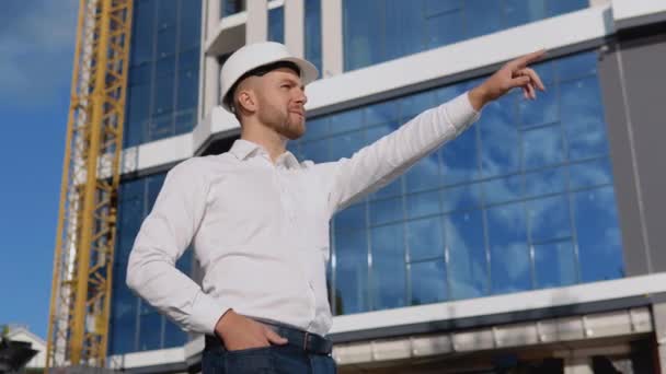 Mérnök építész fehér ingben és sisakban a háttérben egy modern üveg épület kezeli az építési folyamatot — Stock videók