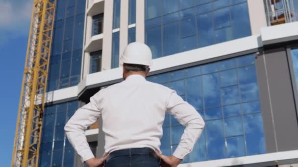 白いシャツとヘルメットの建築家は、現代のガラスの建物を背景にカメラに背を向けて立っています — ストック動画
