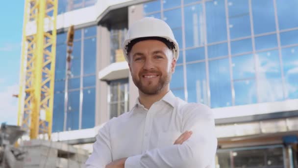 An engineer in a white shirt and helmet stands against the backdrop of a modern glass building. Modern construction — Stock Video