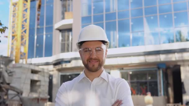 Un ingeniero con camisa blanca y casco se levanta sobre el telón de fondo de un moderno edificio de cristal — Vídeos de Stock