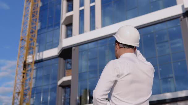 Инженер-архитектор в белой рубашке и шлеме стоит спиной к камере на фоне современного стеклянного здания — стоковое видео