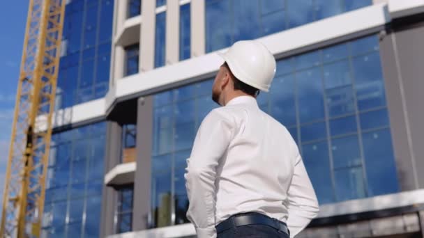 Un architetto ingegnere in camicia bianca e casco sta con le spalle alla macchina fotografica sullo sfondo di un moderno edificio in vetro — Video Stock