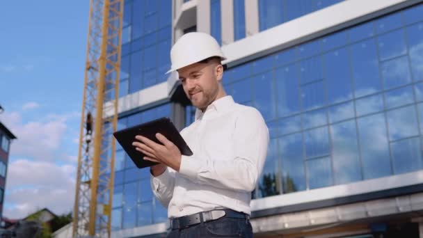 Ingénieur architecte en chemise blanche et casque sur fond de verrerie moderne dans une tablette — Video