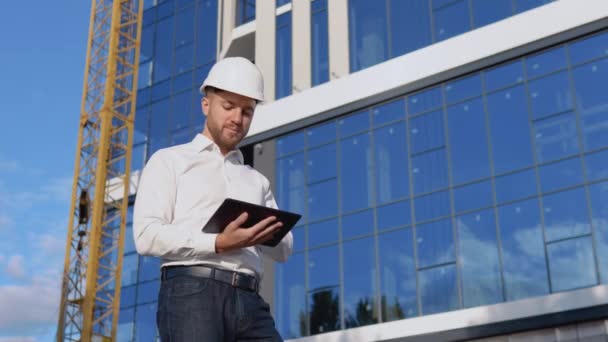 Инженер-архитектор в белой рубашке и шлеме на фоне современного стеклянного здания работает в планшете — стоковое видео