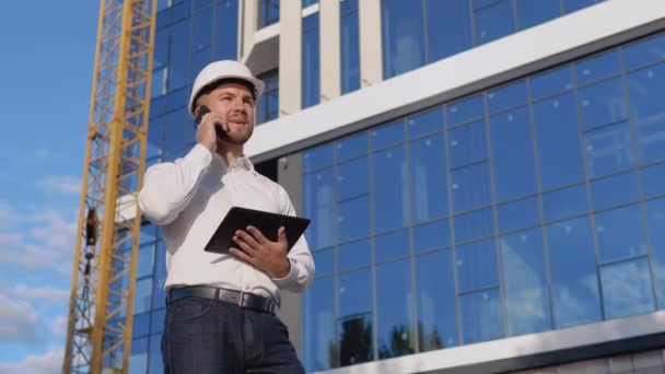 Ein Ingenieur-Architekt in weißem Hemd und Helm vor dem Hintergrund eines modernen Glasgebäudes hält ein Tablet in der Hand und telefoniert mit einem Handy. — Stockvideo