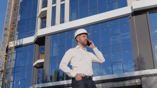 Egy mérnök építész fehér ingben és sisakban egy modern üvegház hátterében mobiltelefonon beszél. — Stock videók