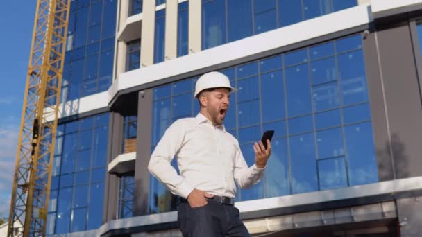 Ingegnere architetto in camicia bianca e casco sullo sfondo di un moderno edificio in vetro parla su un telefono cellulare. Costruzione moderna — Video Stock