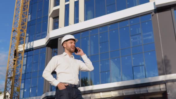 Инженер-архитектор в белой рубашке и шлеме на фоне современного стеклянного здания говорит по мобильному телефону — стоковое видео