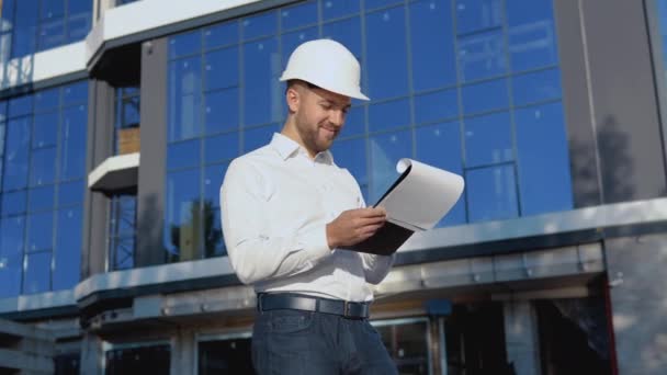 Arquitecto ingeniero en una camisa blanca y casco en el fondo de un edificio de vidrio moderno trabaja haciendo notas en una tableta — Vídeos de Stock