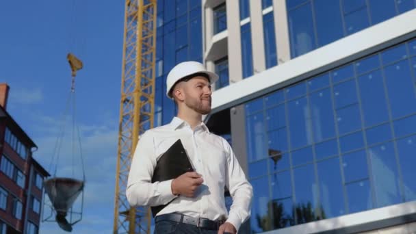 Architect engineer in a white shirt and helmet on a background of a modern glass building holds a tablet — Stock Video