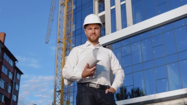 Инженер в белой рубашке и шлеме стоит на фоне современного стеклянного здания и держит рулон с рисунком проекта. Современное строительство — стоковое видео