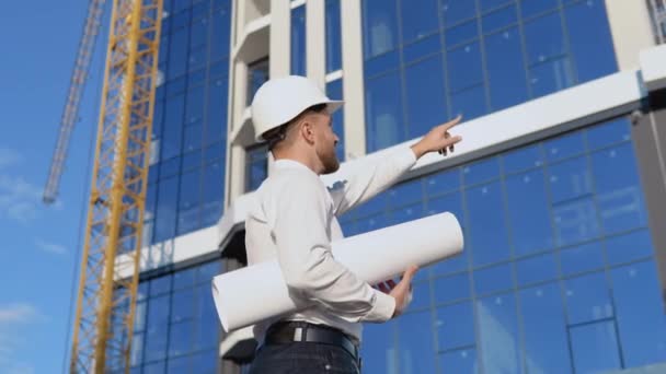 Un ingegnere in camicia bianca e casco lavora alla costruzione di un moderno edificio in vetro e tiene un rotolo con un disegno del progetto — Video Stock