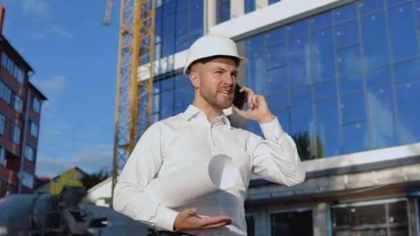Evil engineer in a white shirt and helmet works on the construction of a modern glass building. The engineer-architect holds a roll with a drawing of the project and talks on a cell phone — Stock Video