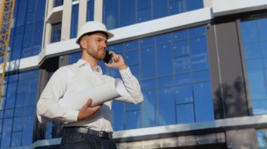 Mühendis-mimar projenin çizimiyle birlikte bir rulo tutuyor ve cep telefonuyla konuşuyor. Beyaz gömlekli ve kasklı bir mühendis modern cam bina inşaatında çalışıyor.