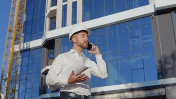 Un ingeniero con camisa blanca y casco trabaja en la construcción de un moderno edificio de cristal. Arquitecto ingeniero lee dibujos de proyectos. — Vídeo de stock