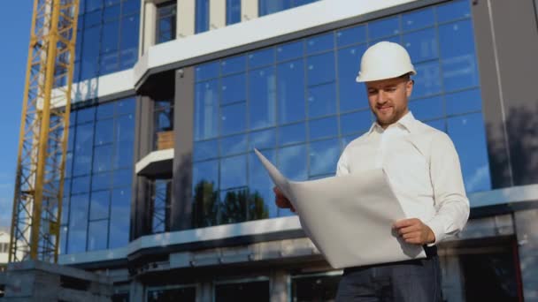 Architekt Ingenieur liest Projektzeichnungen. Ein Ingenieur in weißem Hemd und Helm arbeitet am Bau eines modernen Glasgebäudes — Stockvideo