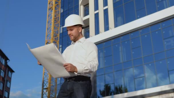 一位身穿白衬衫戴头盔的工程师正在建造一座现代化的玻璃建筑。建筑师工程师阅读工程图纸 — 图库视频影像