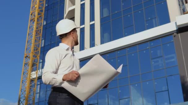 Ingeniero arquitecto lee dibujos de proyectos. Un ingeniero con camisa blanca y casco trabaja en la construcción de un moderno edificio de cristal — Vídeos de Stock