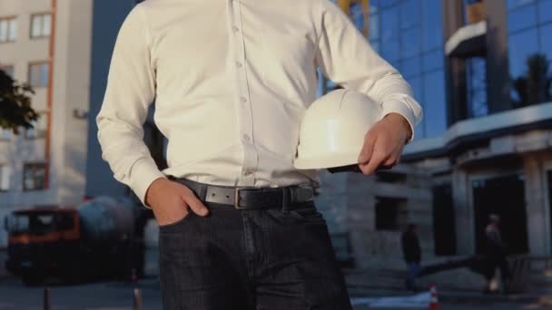 Engenheiro em uma camisa branca no fundo de um edifício de vidro moderno e detém um capacete. Vista de perto de um capacete em uma mão de homem — Vídeo de Stock