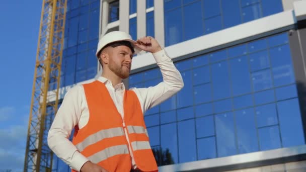 Sullo sfondo di un moderno edificio in vetro si staglia un ingegnere-architetto in camicia bianca, casco e giubbotto da lavoro arancione — Video Stock