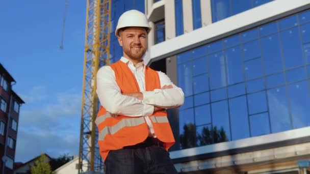 An engineer-architect in a white shirt, helmet and orange work vest stands against the backdrop of a modern glass building — Stock Video