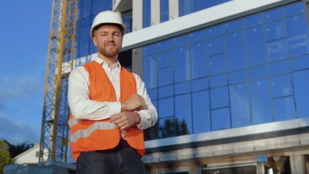 Un ingeniero-arquitecto con camisa blanca, casco y chaleco de trabajo naranja se levanta sobre el telón de fondo de un moderno edificio de cristal — Vídeos de Stock