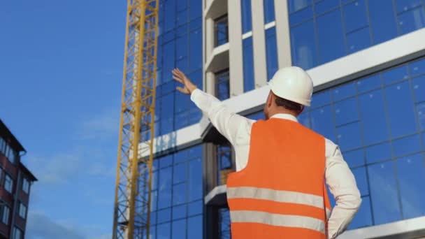 Un ingegnere-architetto in camicia bianca, casco e giubbotto da lavoro arancione si erge con le spalle alla macchina fotografica sullo sfondo di un moderno edificio in vetro e sovrintende al processo di costruzione — Video Stock