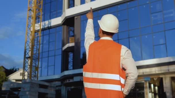 Un ingegnere-architetto in camicia bianca, casco e giubbotto da lavoro arancione si erge con le spalle alla macchina fotografica sullo sfondo di un moderno edificio in vetro e sovrintende al processo di costruzione — Video Stock