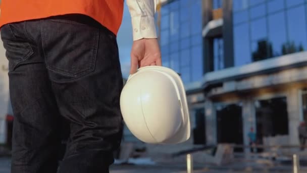 Un ingegnere-architetto in camicia bianca e giubbotto da lavoro arancione cammina in direzione di un moderno edificio in vetro. Vista ravvicinata di un casco nella mano degli architetti — Video Stock