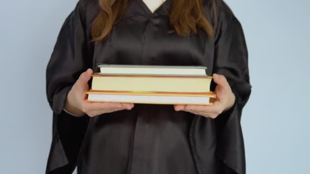 Varios libros de texto en posición horizontal en manos de una estudiante caucásica vestida con una túnica negra. Vista de libros y manos de cerca sobre fondo blanco. — Vídeos de Stock