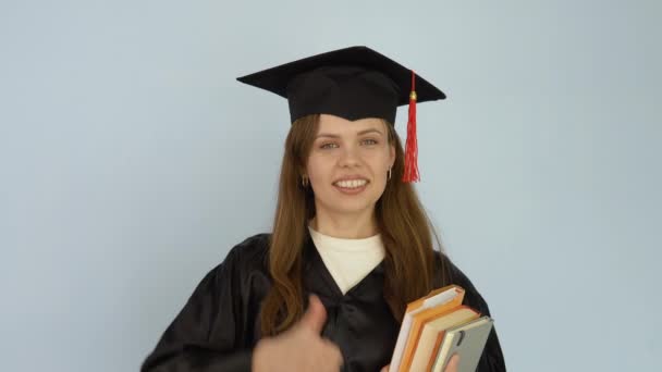 Uma jovem mulher caucasiana em um manto preto e um chapéu mestres fica em linha reta segurando livros didáticos e mostrando o envelhecimento bom com o dedo grande para cima. Estudante olha para a câmera no fundo branco — Vídeo de Stock