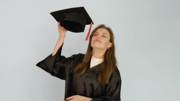 Uma jovem estudante caucasiana vestida com um manto preto brinca com um chapéu quadrado e faz cócegas no nariz com uma escova. Fundo branco — Vídeo de Stock