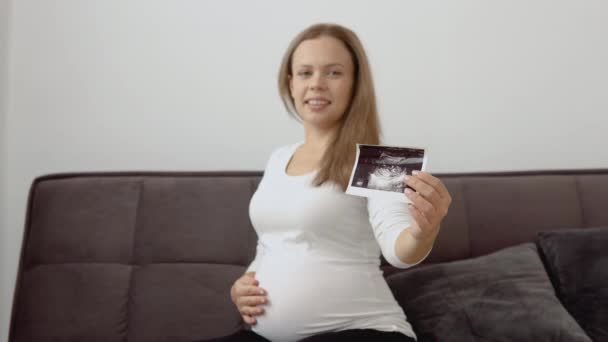 Hamile, açık tenli bir kadın karnını okşar ve ceninin ultrason teşhisinin fotoğrafını gösterir. — Stok video