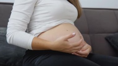 Hamile, açık tenli bir kadın rahat bir koltukta oturuyor ve karnını okşuyor.