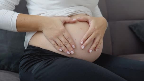 Une femme enceinte s'assoit sur un canapé confortable et fait un signe de coeur sur son ventre avec ses mains — Video