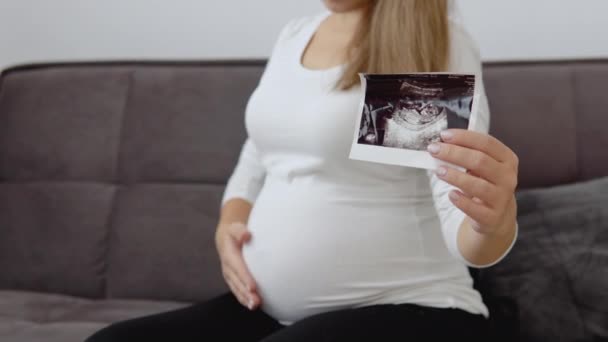 Uma mulher grávida de pele clara acaricia a barriga e mostra uma foto do diagnóstico de ultra-som do feto — Vídeo de Stock