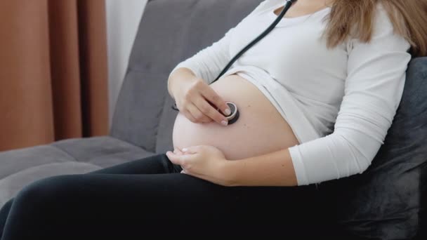 Una mujer embarazada escucha los movimientos y los latidos del corazón del feto con un estetoscopio.. — Vídeos de Stock