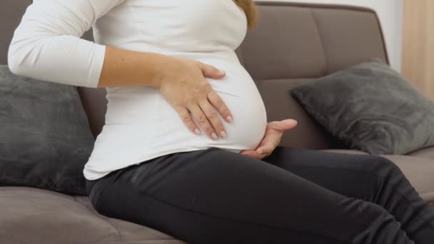 Těhotná žena sedí na pohodlné pohovce a hladí si bříško — Stock video
